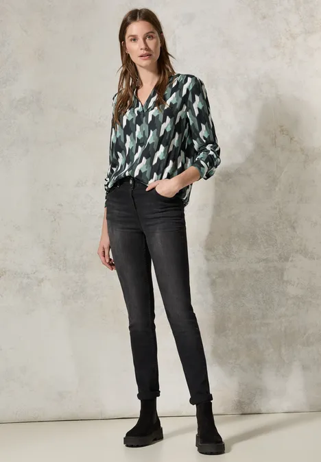 Frau mit gemusterter Bluse, schwarzer Jeans und Stiefeln vor hellem Hintergrund.